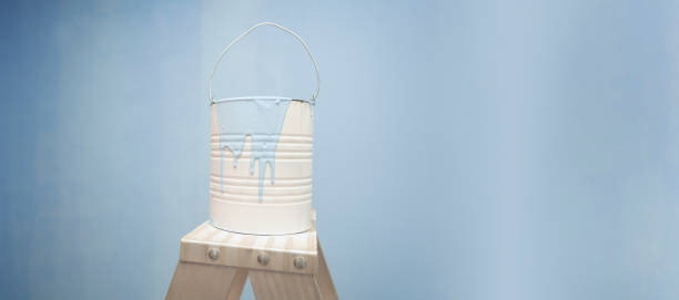 white paint jar with blue paint dripping on a metal ladder, with a blue wall background. Image with copy space