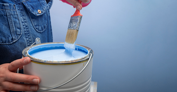 Close up on woman wearing overalls taking out the brush from the jar of blue paint next to a blue wall