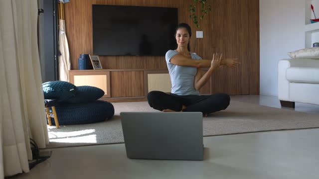 Beautiful woman following an online class doing some warm up exercises using a laptop and sitting on the floor at home