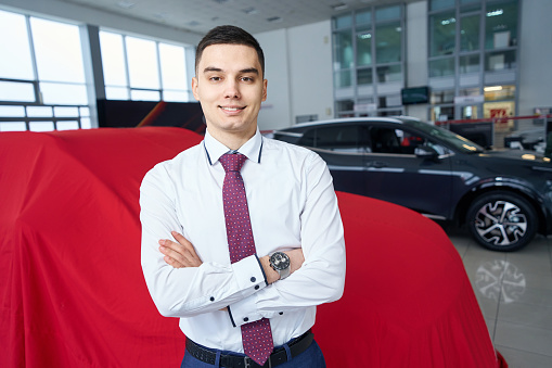 Smiling young man stands in front of a car covered with red cloth, the manager is in modern car dealership