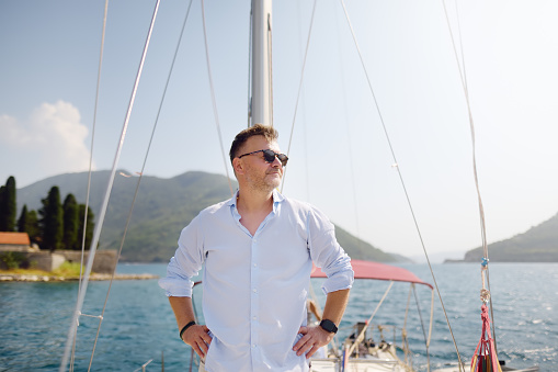 Handsome mature man aboard sailboat in Bay of Kotor near town of Perast on Adriatic Sea in Balkan Mountains, Montenegro, Europe. Happy person on sea voyage on sunny day of summer vacation.