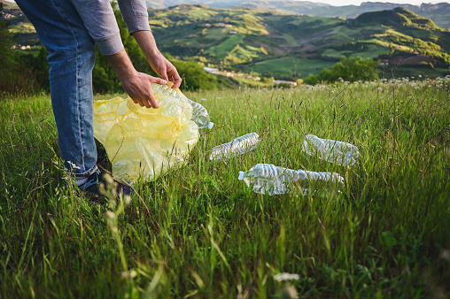unrecognizable man picking up plastic bottle in forest meadow in mountains.