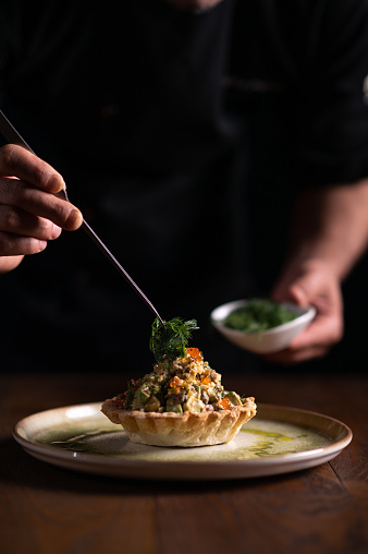 A male chef decorating a plate of tartare di pesce spada with a pinch of dill on a dark background.