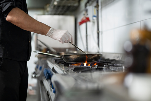 A male chef preparing food over a flaming gas stove with pan in restaurant kitchen.