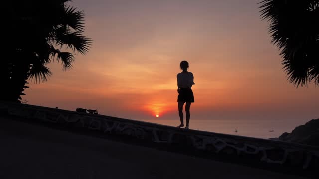 Woman is jumping from the curb standing on the coastline at summer evening against sunset over sea
