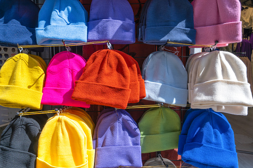 Lots of brightly colored knitted beanie hats hanging on hangers in spontaneous cheap street market. Hats on clothespins hang in window low-quality clothes fake. Rows of cap showcase sale collection
