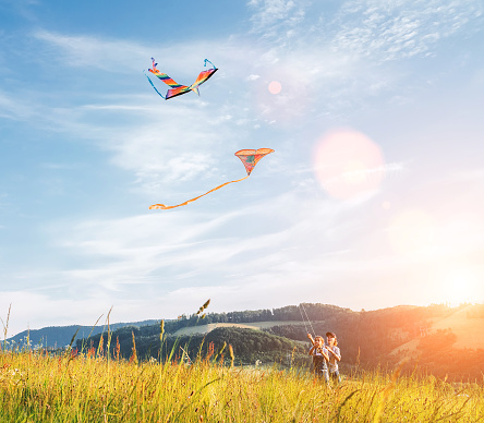 Smiling sister with launching brother with colorful kites - popular outdoor toy on the high grass hills meadow. Happy childhood moments or outdoor time spending concept image.
