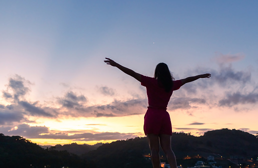 Silhouette of a young woman with arms raised and the sky after sunset in the background.