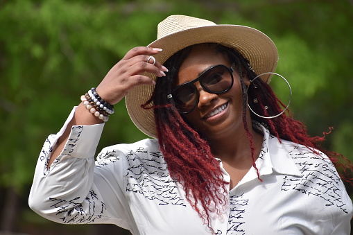 4-19-2023 Houston, Texas - Exceptionally beautiful plus-size model of color smiling on a bridge with designer shades and a summer hat; forest trees in the background