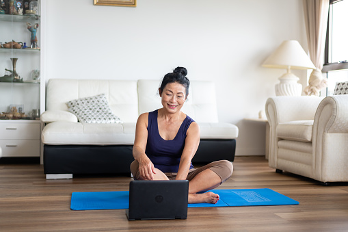 A mature aged woman of asian ethnicity works out from the comfort of her living room. She is wearing casual active wear and smiling as she selects the class she would like to attend off her laptop.
