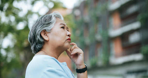 Portrait of thoughtful mature woman with gray hair standing and looking away, Anxiety appeared on her face, modern building in background, copy space to insert your advertisement