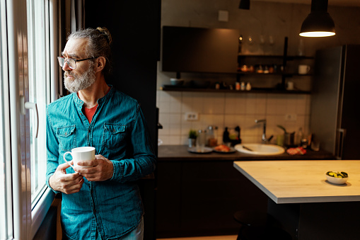 One mature man looking through window while drinking morning coffee at home