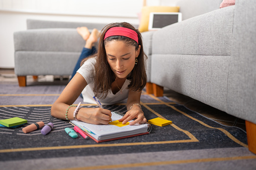 Young female student with pink headband, lying on the carpet in the living room of her apartment, writing in her notebook
