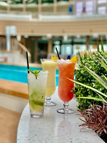 A frozen red cocktail (could be a mai tai, or a hurricane, a rum runner or a strawberry daiquiri) garnished with an orange slice sits on a lake side wooden table. The vibrant greens and blues of the water in the background.