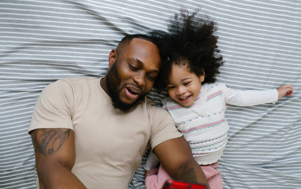 Top view of cheerful bearded black father and his little daughter taking selfie while lying on bed in beddrom. Happy family African American moments, relax cozy leisure lifestyle together at home.