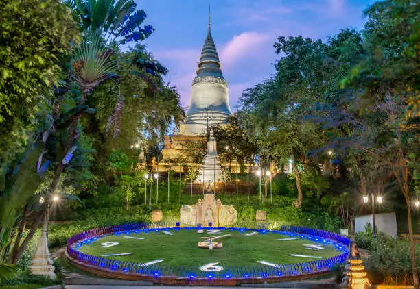 In Wat Penh gardens,a gift from China,covered in green grass,a large working clock,beneath the hilltop pagoda of Wat Phnom,a prominent city landmark,next to the circular pathway.