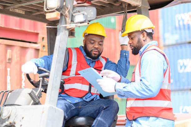 istock Two African American males in safety uniforms and helmets, container operating working with the forklift truck driver at commercial dock site. Cargo Ship Import Export Factory Logistic.. 1485312734