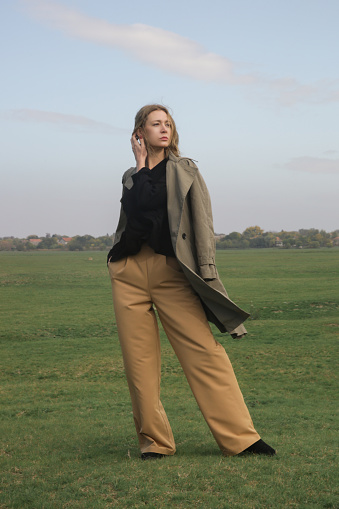 Outdoor fashion portrait of Caucasian woman in wide leg beige pants, black blouse and classic green trench coat