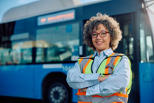 Happy female bus driver standing with crossed arms at the station and looking at camera.