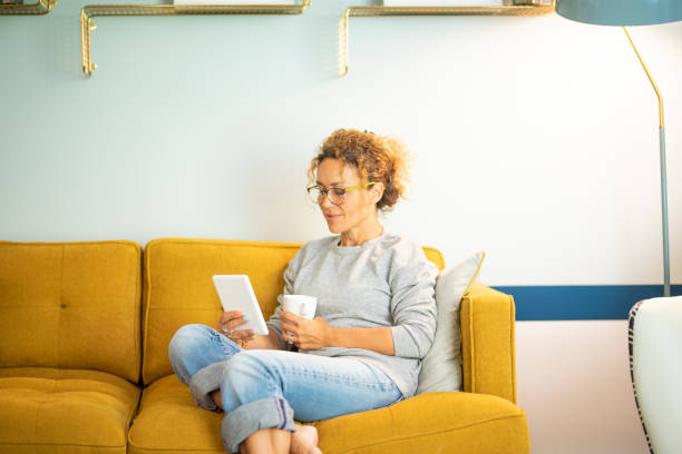 istock Smart casual woman middle age relaxing at home sitting on yellow sofa and using ereader to read an ebook enjoying indoor leisure activity alone. Sunday time lifestyle. People and technology online 1485330026