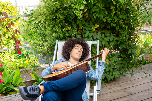 Beautiful young Afro-Italian mixed race man in casual wear playing guitar in the garden. A multiracial boy plays a musical instrument rests and relaxes outdoors. Spending time at home