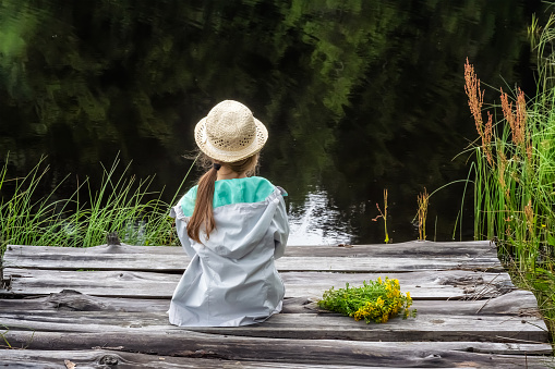 Beautiful little girl with a hat, taking a rest while sitting on a wooden pier of a mountain lake holding a bouquet of herbs