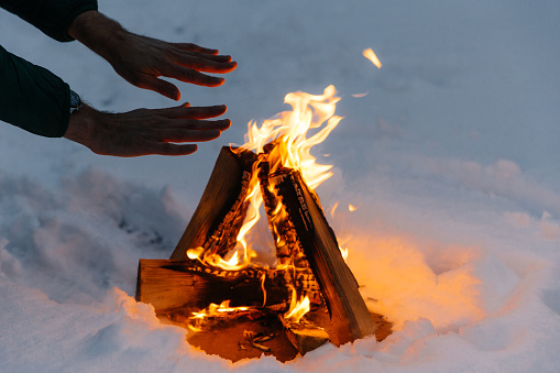 Unrecognizable male warms hands on fire in forest during cold winter, tries to warm himself, being cold spend time on cold snow and frosty weather. Burning flame on ground covered with snow.