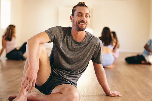 Happy young man sitting in a yoga studio with a group of people in the background. Smiling young man having a yoga session with his class in a community fitness center.