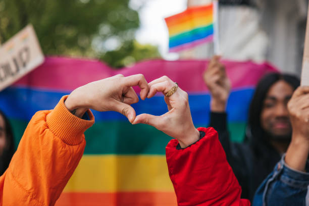 Genderqueer and non-binary friends making a heart shape against a rainbow flag Genderqueer and non-binary friends making a heart shape against a rainbow flag celebrating love during a Pride Parade. pride stock pictures, royalty-free photos & images