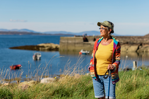 A wide-view shot of a senior woman standing by the bay near the village of Diabaig on the side of Loch Torridon in Wester Ross, Scotland. She is using a hiking pole and enjoying the nice weather.