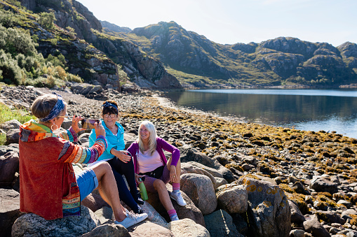 A shot of a small group of senior women sitting down at the bay on the rocks near the village of Diabaig on the side of Loch Torridon in Wester Ross, Scotland. They are taking a break from their hike to sit, relax and chat, while one woman takes a picture of her friends with a smartphone. One woman is holding a reusable water bottle.