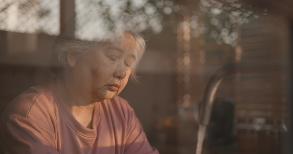 View through glass window shot, Asian senior woman absentmindedly feel sadness, face expression sorrow while look out of the window of kitchen during she washed a dishes in her hand