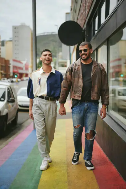 Smiling young gay couple walking hand in hand together along a city sidewalk painted with the pride rainbow