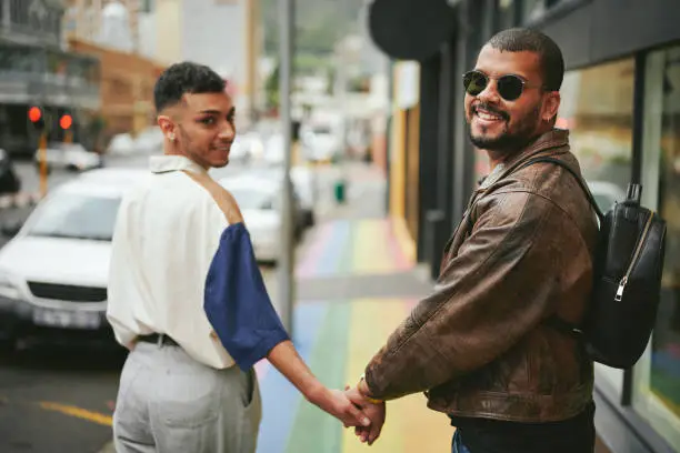 Portrait of a young gay couple looking over their shoulder and smiling while walking together on a sidewalk painted with the pride rainbow