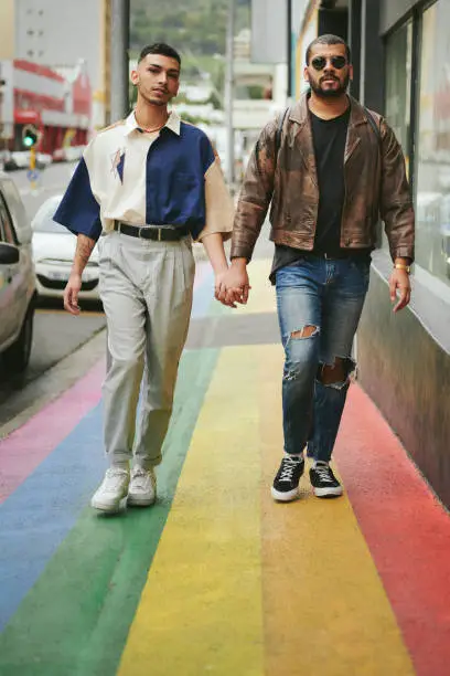 Young gay couple walking hand in hand together along a city sidewalk painted with the pride rainbow