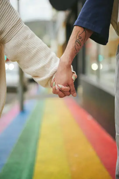 Close-up of two young friends standing hand in hand in unity on a city sidewalk painted with the pride rainbow