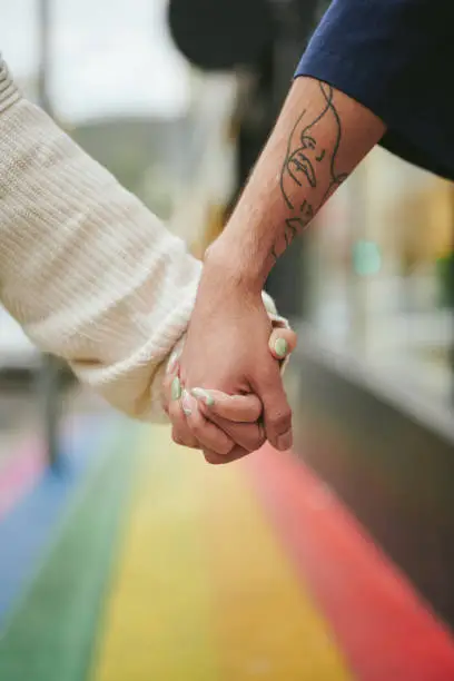 Close-up of two young friends holding hands in unity together on a city sidewalk painted with the pride rainbow