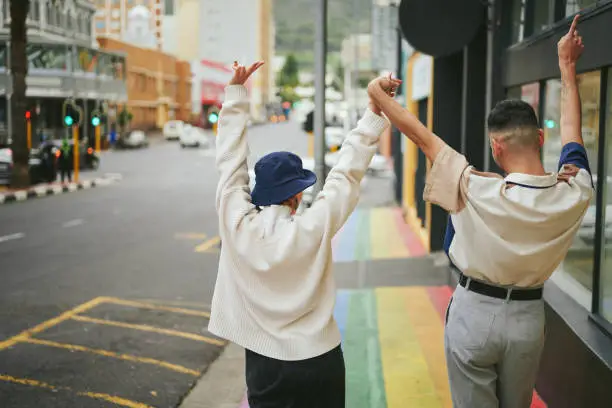 Rear view of two carefree young friends dancing together on a city sidewalk painted with the pride rainbow