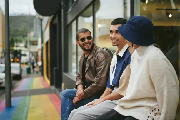 Group of smiling young friends talking while sitting in front of a store on a sidewalk painted with the pride rainbow