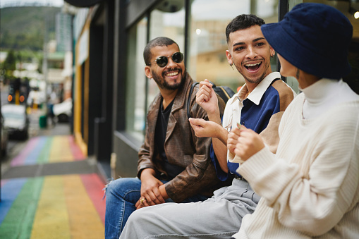 Laughing group of young friends talking while sitting in front of a store on a sidewalk painted with the pride rainbow