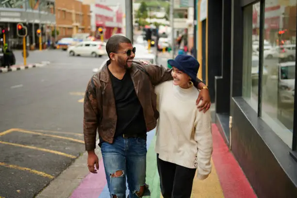 Two young friends talking together and laughing while walking arm in arm along a city sidewalk painted with the rainbow flag