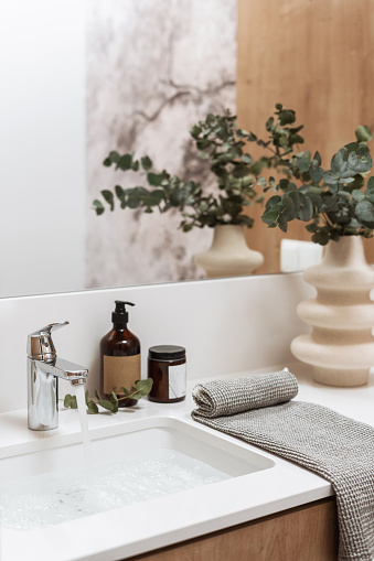 Interior with washbasin filled water, metal faucet, vanity mirror, soap dispenser, jar with face cream, rolled towel and ceramic vase with green branch of eucalyptus in bathroom