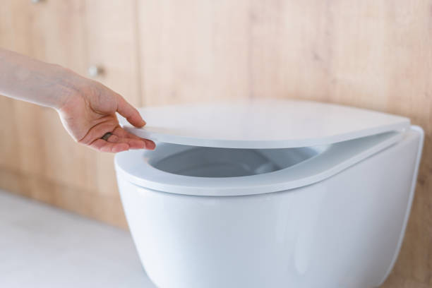 Woman closing the lid of a toilet seat Woman hand closing the lid on toilet seat in the modern bathroom or wc. Plumbing system at restroom installed on wall with wooden panels. Hygiene and health care concept toilet bowl stock pictures, royalty-free photos & images