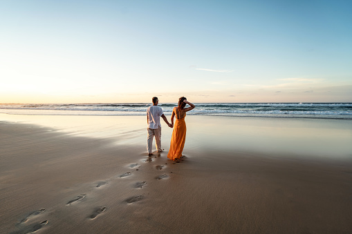 Romantic couple walking on sunset beach, enjoying evening light, relaxing on tropical summer vacation. Honeymoon. Love. Back view. Woman wearing orange maxi dress, man in linen pant and white shirt.