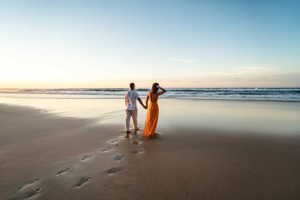 casal romântico caminhando na praia do pôr do sol, desfrutando da luz da noite, relaxando nas férias tropicais de verão. lua de mel. amor. vista traseira. - honeymoon wedding beach couple - fotografias e filmes do acervo