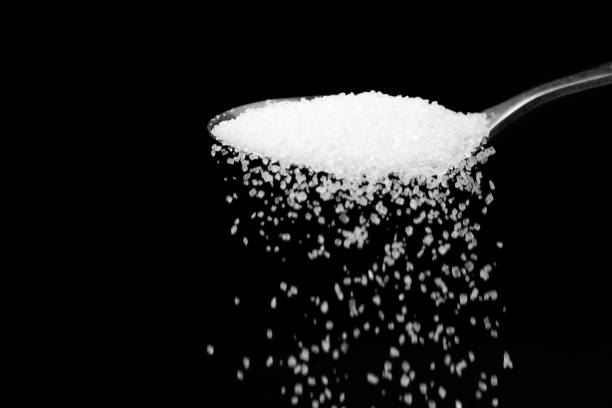 Macro photography of a teaspoon with crumbling white sugar on a black background, copy space.