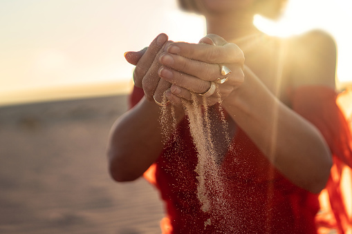 Elegant woman with sand falling through her hands on the beach at sunset time. Golden light. Conceptual photo.