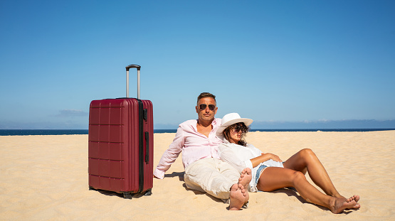 Traveling couple relaxing with suitcases at the beach on a sunny summer day, tired after looking for the good place for sunbathing. A lot of copy space. Tourism.