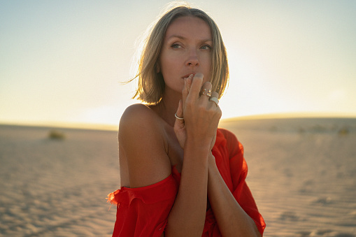 Portrait of a romantic blonde woman in a red dress posing in the sand desert at golden sunset light.