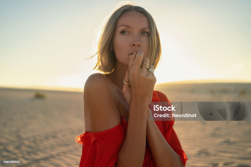 Portrait of a romantic blonde woman in a red dress posing in the sand desert at golden sunset light. Portrait of a romantic blonde woman in a red dress posing in the sand desert at golden sunset light. Girl wearing fashionable rings on hands. Women Stock Photo